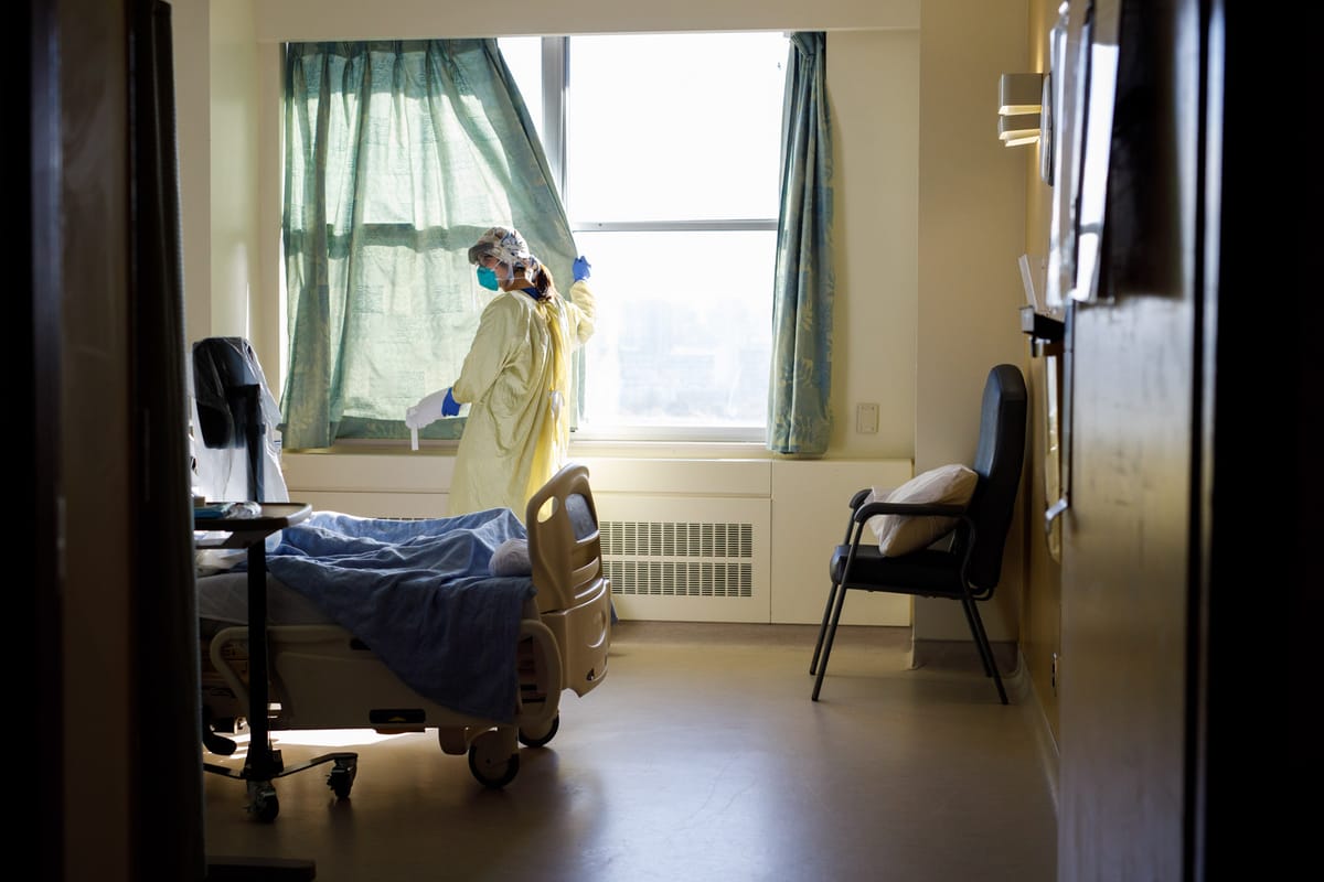 A wide-angle photo of a healthcare worker in a yellow hospital gown next to a hospital bed, drawing the curtains closed in a patient room
